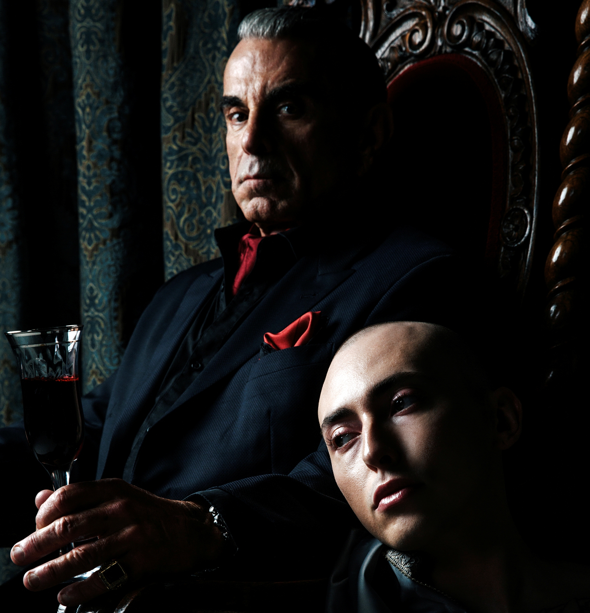 vampire in nice suit, red cravat, red pocket square, glass of wine on a throne with a bald servant at his knee